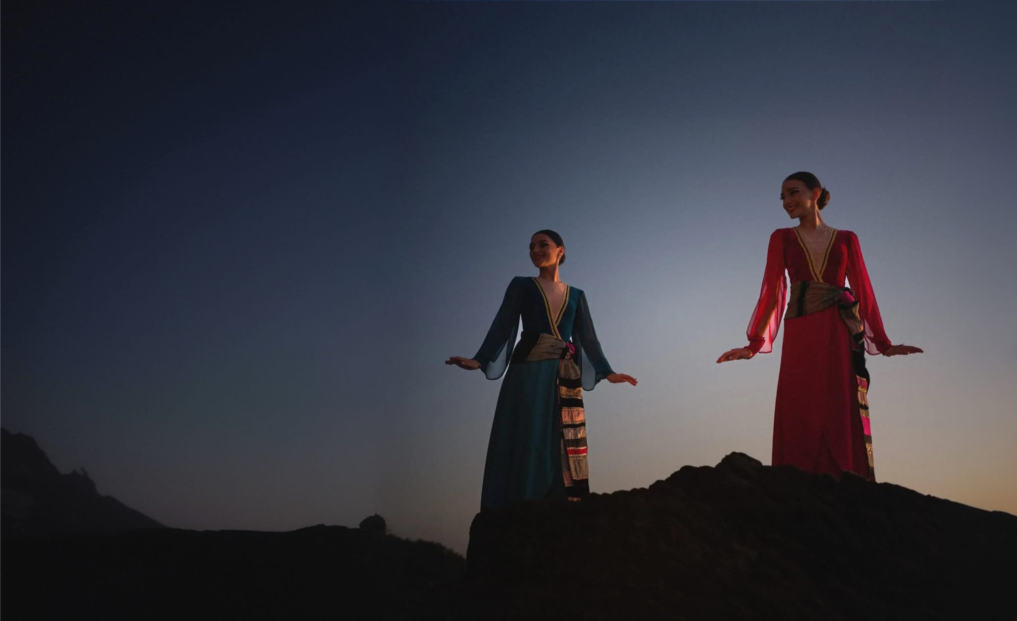Two dancers in traditional dresses on a mountain ridge at dusk